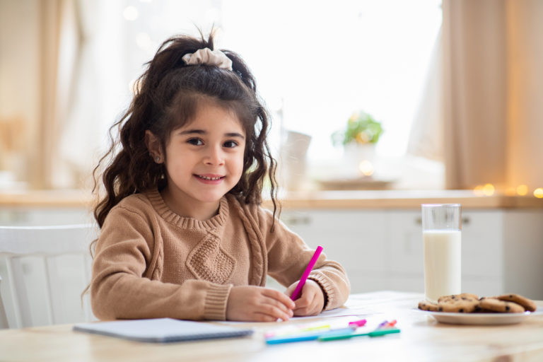 Children Development. Adorable Preschool Arab Girl Drawing In Kitchen At Home And Smiling At Camera, Cute Little Kid Sitting At Table And Painting With Colorful Pencils, Free Space